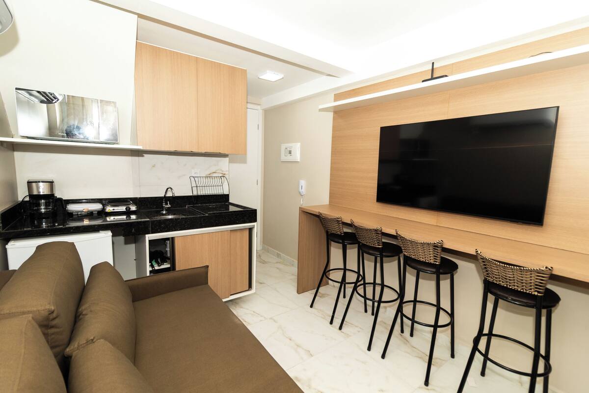 A modern kitchen with a built-in stove, a coffee machine, and a small refrigerator. There is a bar area with stools and a flat-screen TV mounted on the wall.