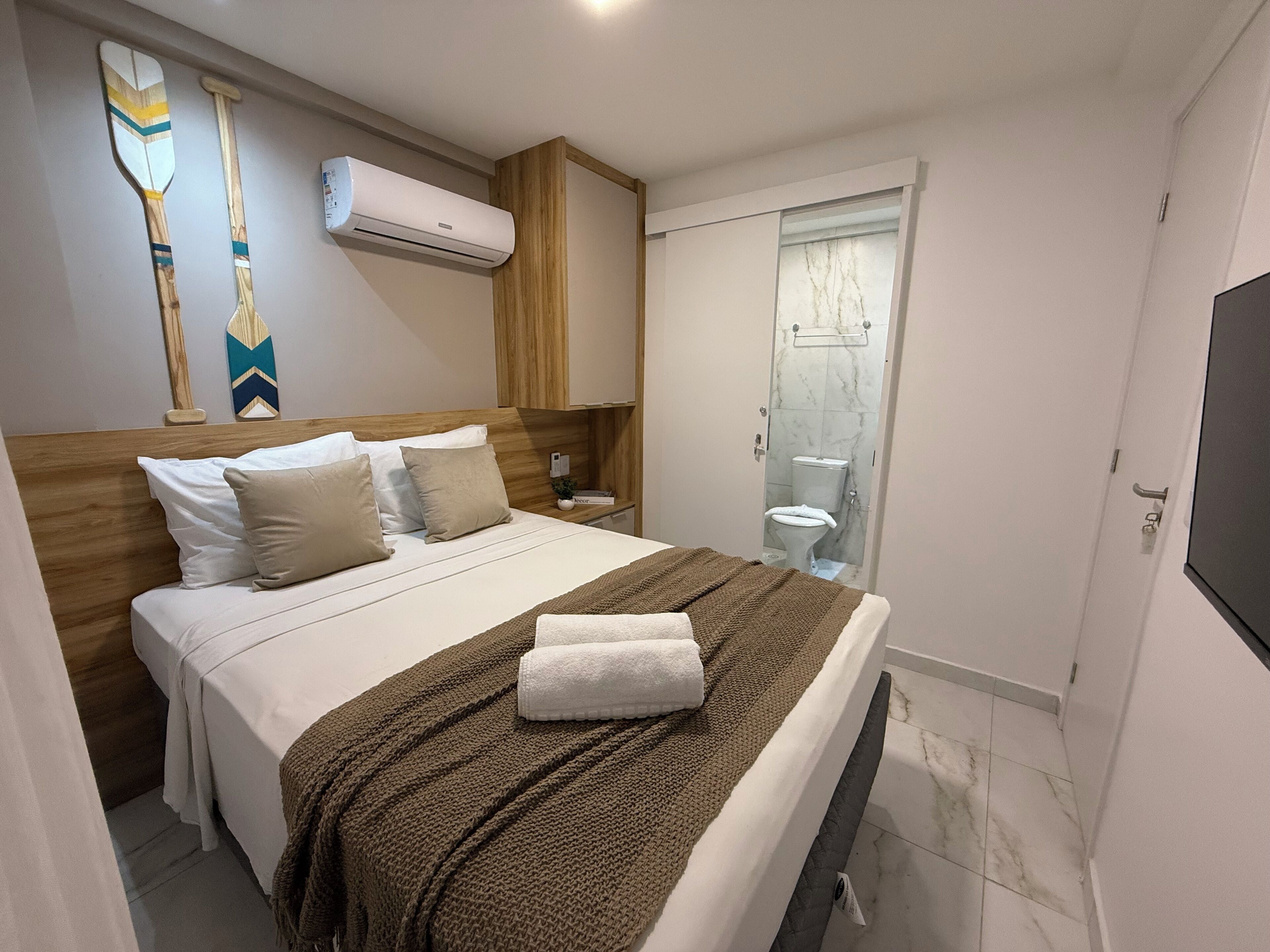 A neatly made bed with a brown blanket, a wooden headboard, and a wall-mounted air conditioner.
