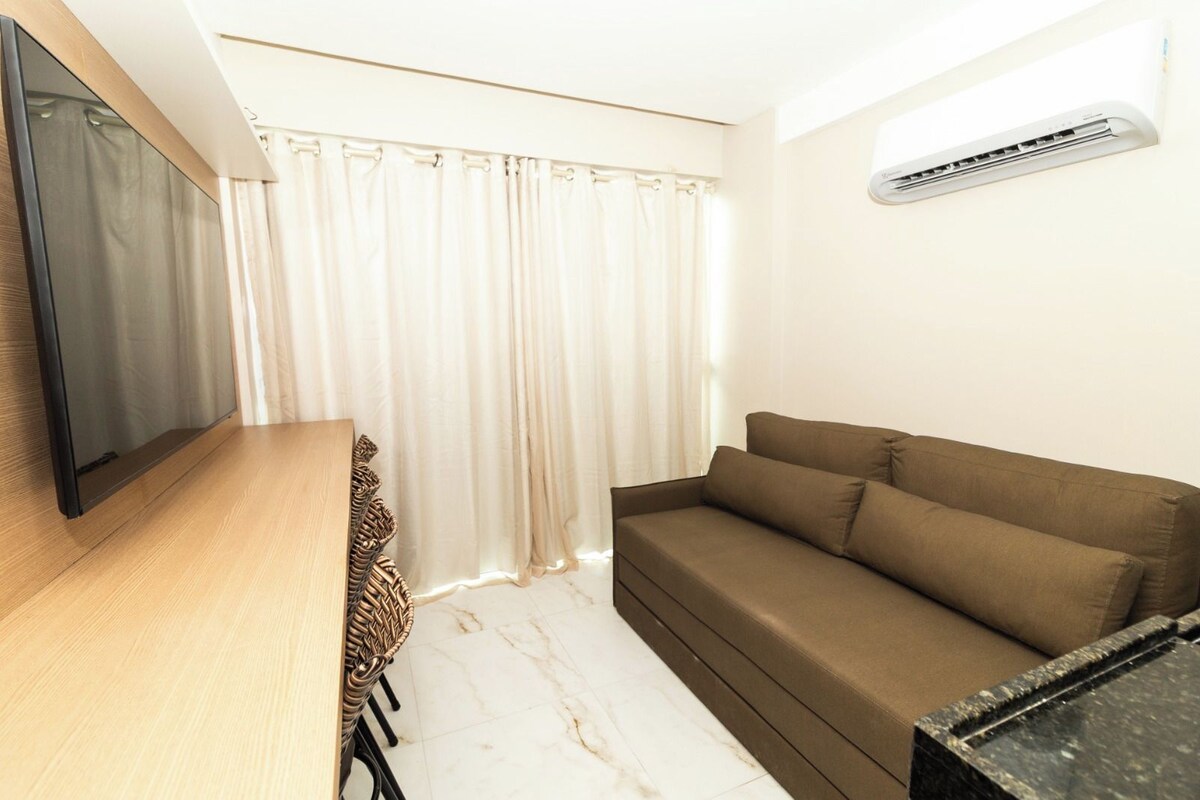 A room with a flat-screen TV, a brown sofa, a wooden table, and a white curtain.