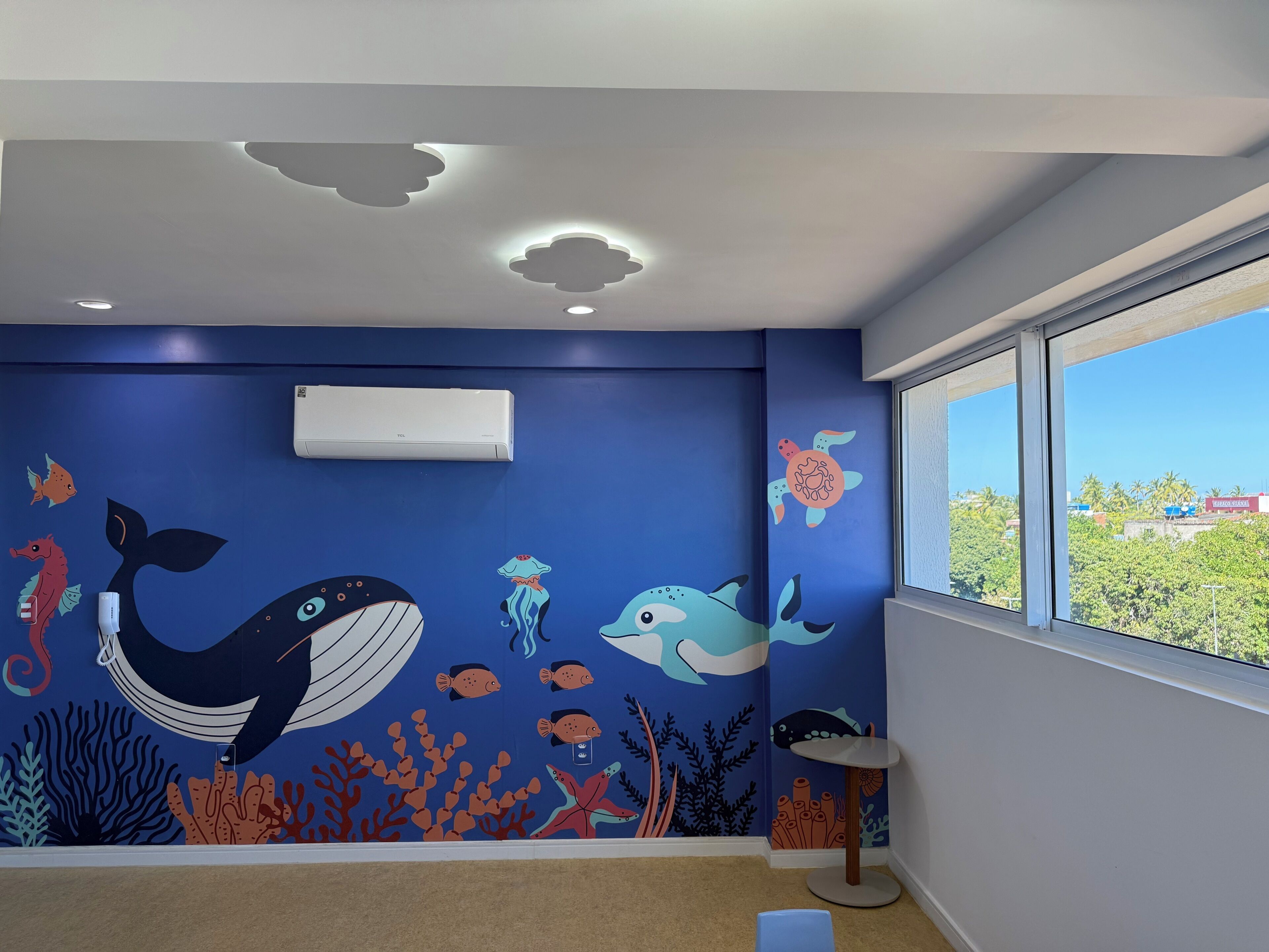 A room with a blue wall featuring an underwater mural, a white air conditioning unit, a round table, and a window with a view of greenery.