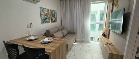 Una sala de estar moderna con una mesa de comedor de madera, dos sillas negras, un sofá beige y un televisor montado en la pared.