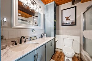Ein Badezimmer mit Toilette in Weiß, einem Waschbecken mit Marmorplatte, einem Spiegel mit Beleuchtung darüber und einem gerahmten Bild von einem Hund in einer Badewanne.