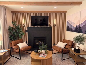 Uma sala de estar moderna com duas poltronas de couro, uma mesa de centro de madeira, uma lareira e uma televisão grande fixada acima.