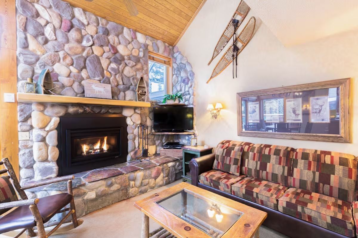 A cozy living room with a stone fireplace, a television, a patterned sofa, and a glass coffee table.