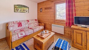 Interior acollidor d'una cabana amb un sofà de fusta, una taula petita de fusta amb una safata, una televisió i una finestra amb cortines.