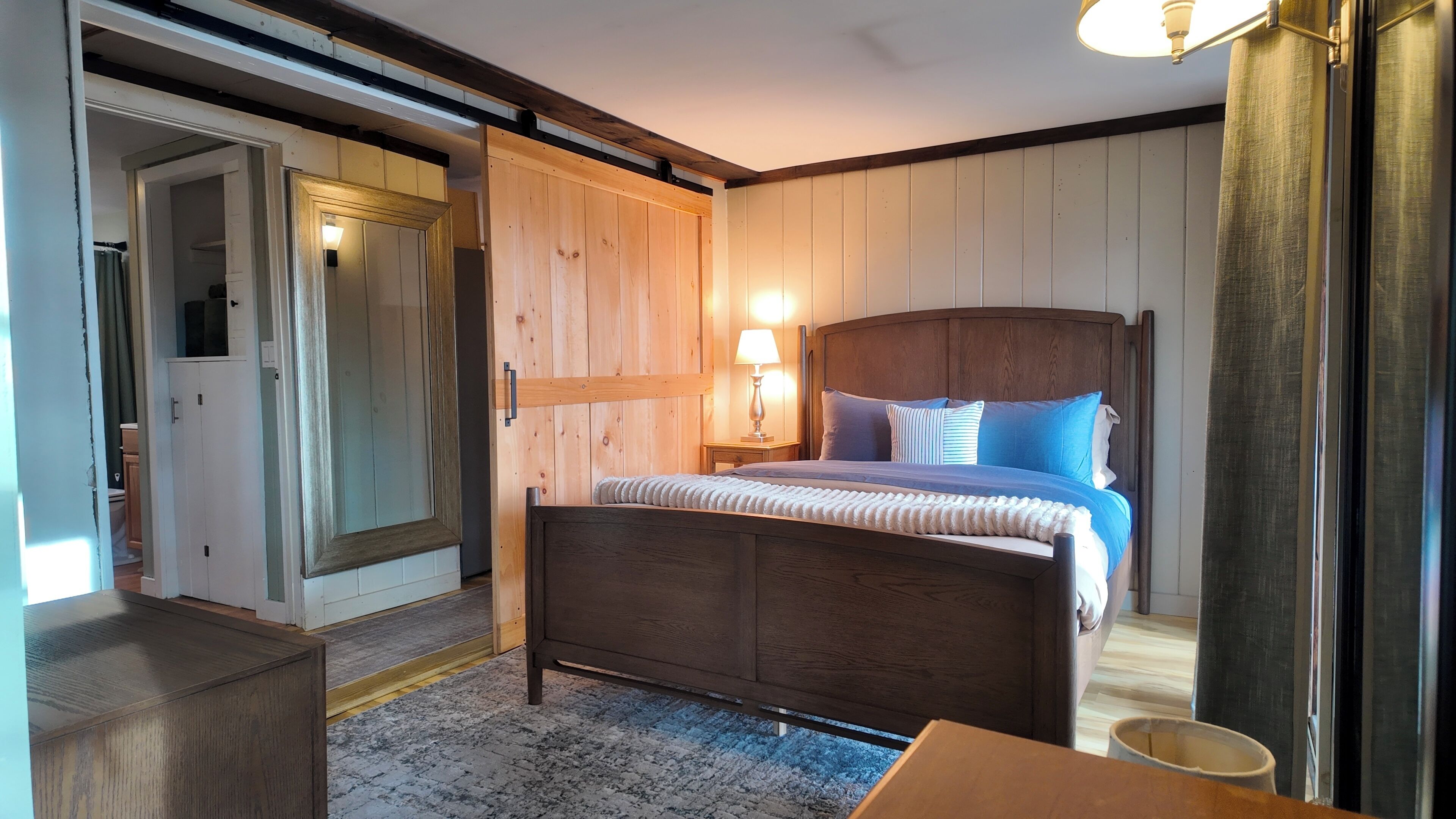 A bedroom with a wooden bed, a nightstand with a lamp, a wardrobe, and a door leading to another room.