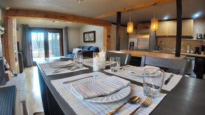 A dining area with a long table set for four, featuring a modern kitchen in the background with wooden accents and stainless steel appliances. - Peaceful Family Friendly Private Lake Waterfront Home Close to Beach and Hiking (Bennington)