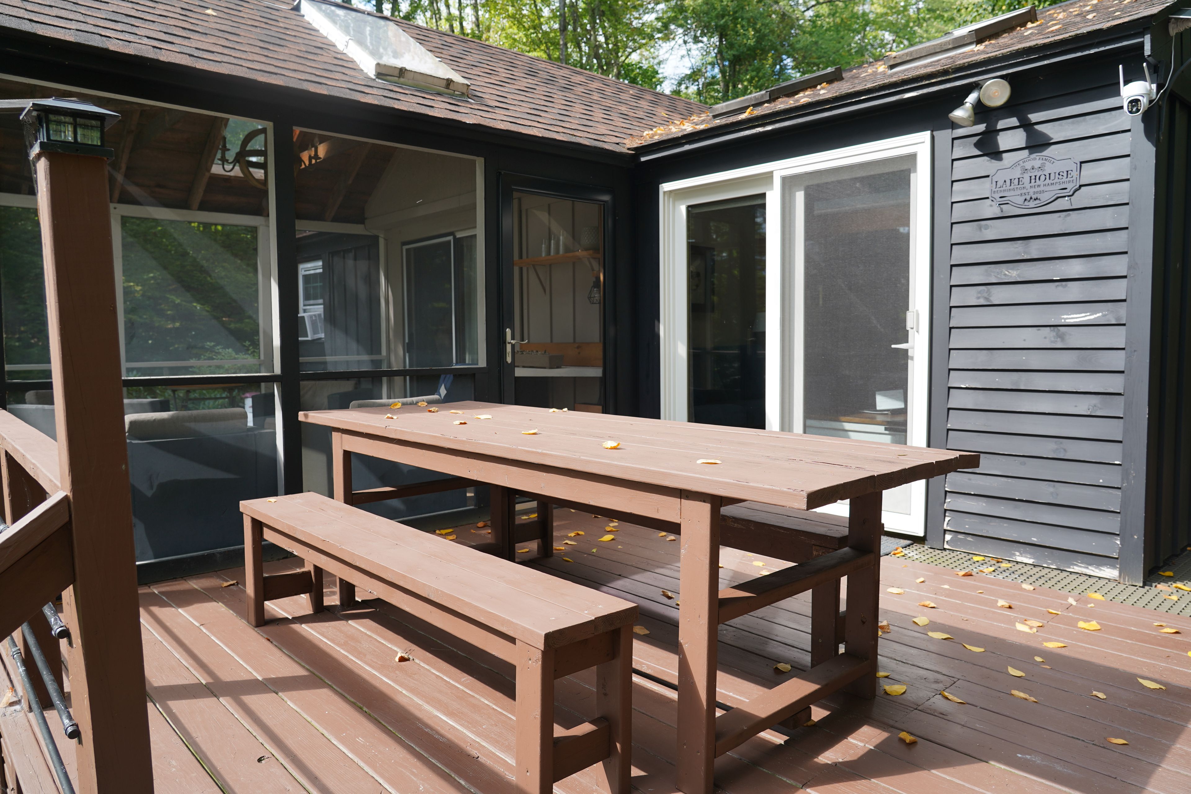 Une terrasse en bois avec une table de pique-nique et un banc, un patio couvert et un bâtiment sur lequel est affiché un panneau indiquant « MAISON AU BORD DU LAC ».