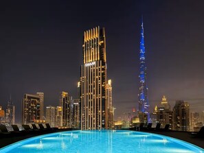 A rooftop pool with a city skyline in the background, including a prominent building with vertical lights and a tall tower with a spiral design. - Stayshort - Lovely 3 Bed in Downtown Sleeps 5 With Balcony (Dubai)