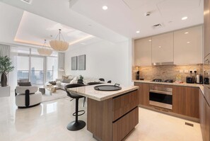 A modern kitchen with a central island, wooden cabinets, and stainless steel appliances. There is a dining area with a sofa and a coffee table. - Stayshort - Lovely 3 Bed in Downtown Sleeps 5 With Balcony (Dubai)