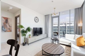 A modern living room with a flat-screen TV, a sofa, a dining table, and large windows offering a city view. - Stayshort - 1 Bed Unit in Meydan Sleeps 2 With Balcony (Dubai)