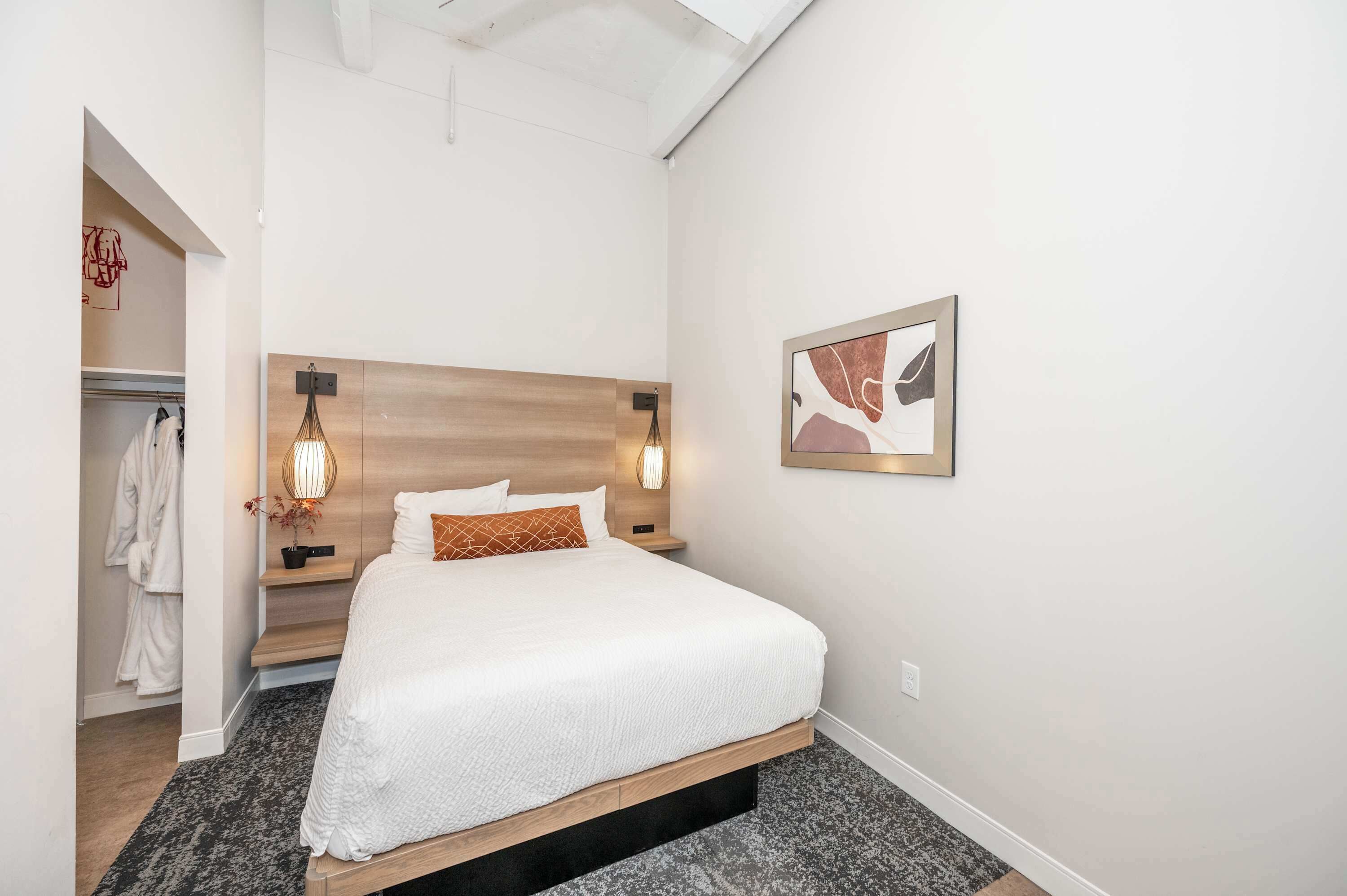A modern hotel room with a large bed, bedside lamps, a wooden headboard, a framed abstract artwork, and a closet with a robe hanging.