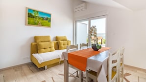 A dining area with a table set for four, a sofa with cushions, a painting on the wall, and a view through a window. - Stunning apartment in Peroj with house a panoramic view (Peroj)