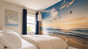 Ein Schlafzimmer mit einer großen Wandmalerei eines Sonnenuntergangs über dem Meer, einem Bett mit weißer Bettwäsche, einer Karte an der Wand und einem Fenster mit Vorhängen.