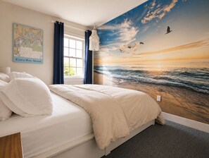 A bedroom with a large mural of a sunset over the ocean, a bed with white bedding, a map on the wall, and a window with curtains. - Charming Coastal Suite (Freeport)