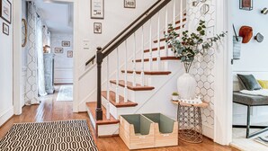 Un escalier avec des marches en bois et des rampes blanches, menant à une pièce où se trouvent un tapis à motifs, un vase avec des plantes et une petite table sur laquelle est posé un objet décoratif.