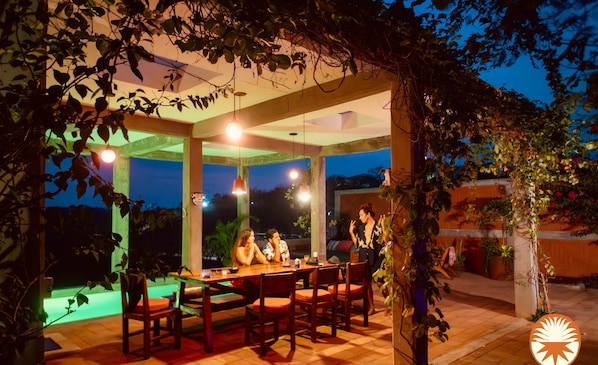 An outdoor dining area with a covered table and chairs, surrounded by plants and a view of the night sky.