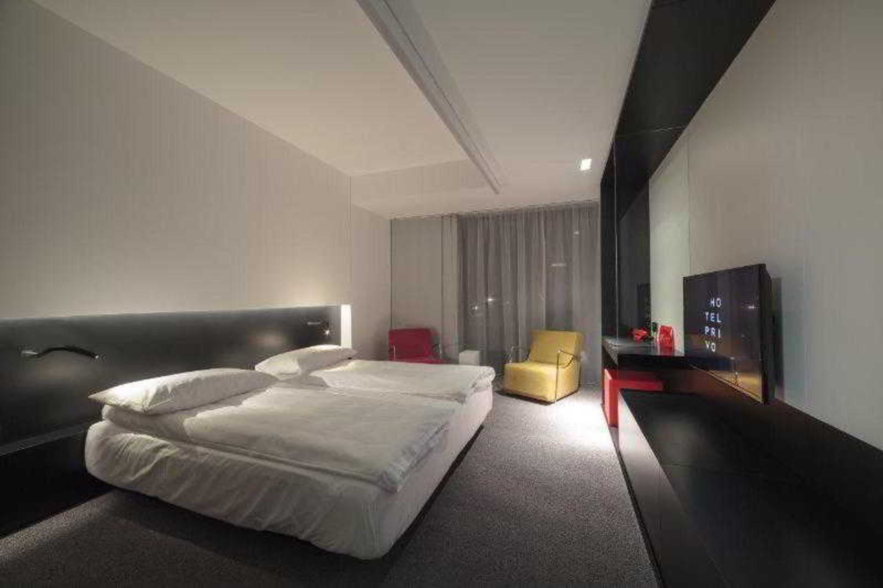 A modern hotel room with a large bed, a small armchair, a TV, and a mirror.