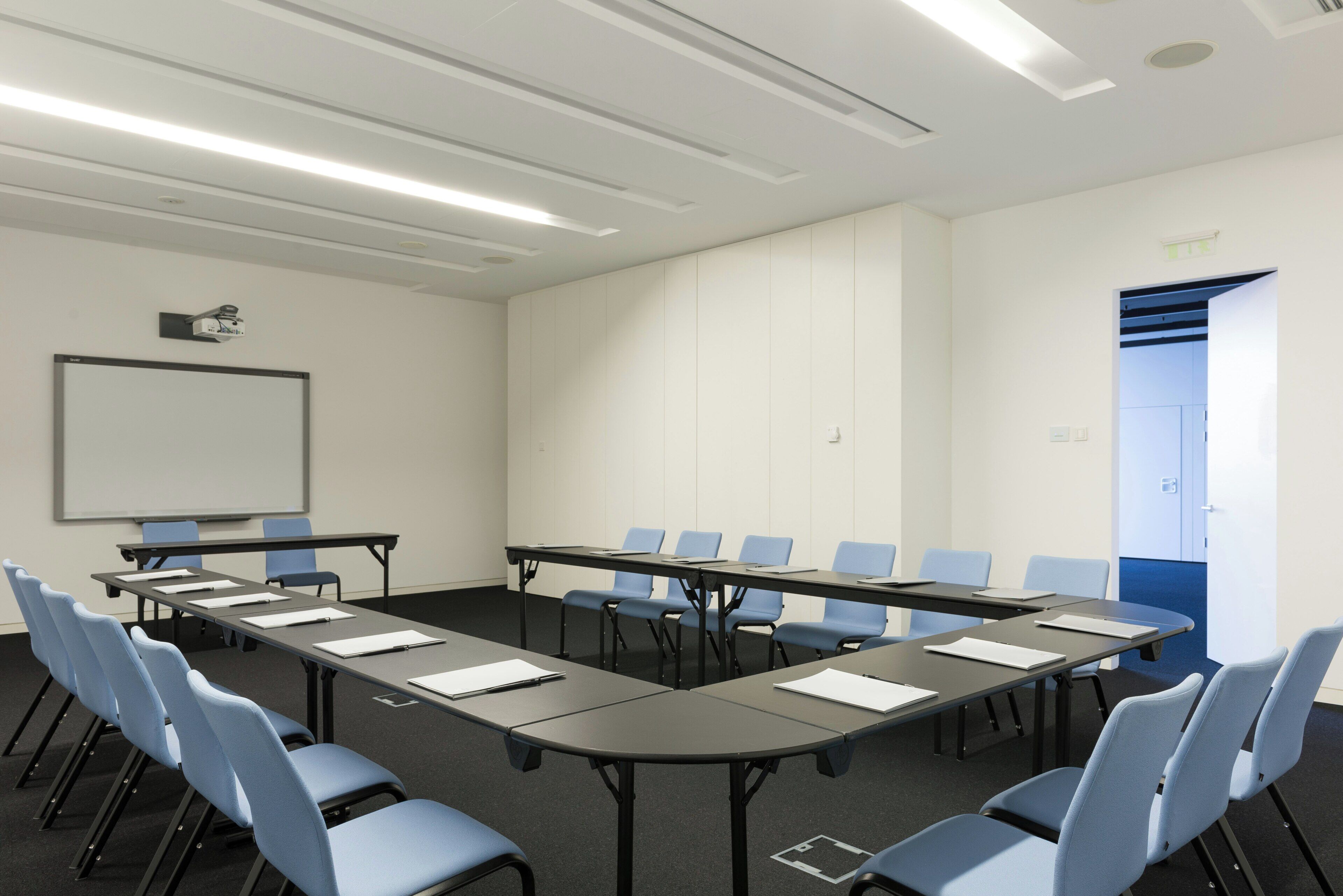 Uma sala de conferências com uma mesa comprida, cadeiras azuis, uma tela de projetor e um quadro branco.