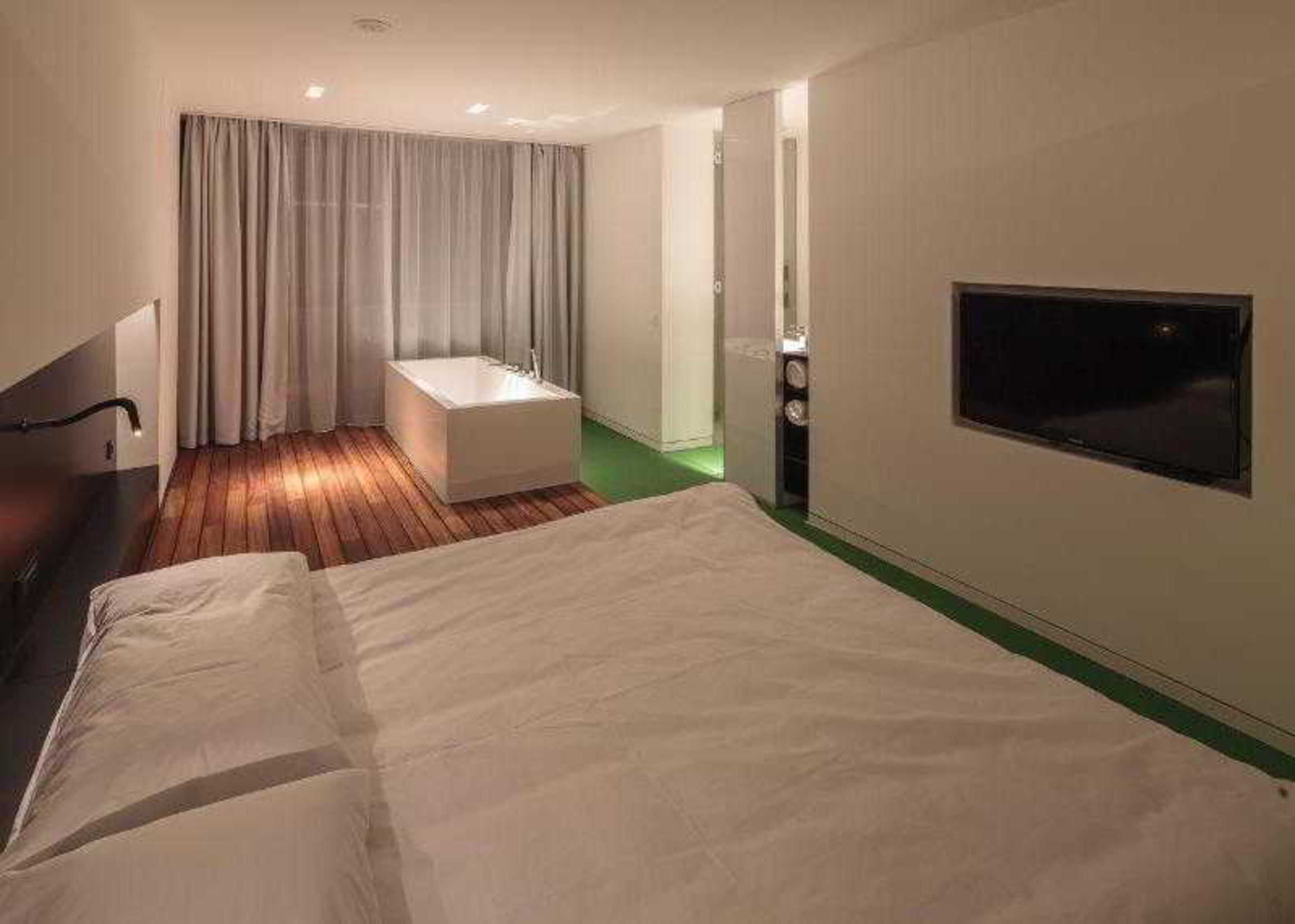 A hotel room with a bed, a small sitting area with a bench, a TV, and a shower area.