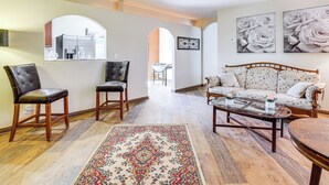Un saló amb un sofà, dues cadires, una taula baixa i una catifes. Es pot veure una cuina a través d'una arcada.