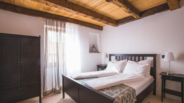 Ein Schlafzimmer mit Holzbalkendecke, einem Bett mit weißer Bettwäsche, einem Nachttisch mit Lampe und einem Kleiderschrank.