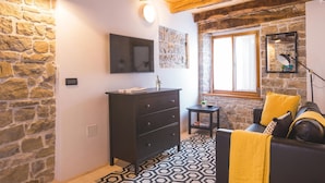 Una habitación con pared de piedra, un televisor de pantalla plana, un aparador de madera oscura, una pequeña mesa con una planta y un plaid amarillo sobre un sofá negro.