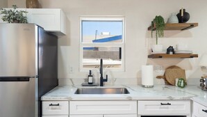 Una cocina con un refrigerador de acero inoxidable, un lavamanos con grifo, una ventana con vista a un edificio, y estantes con vajilla y plantas.