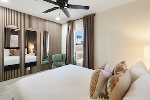 A hotel room with a bed, a ceiling fan, a window with curtains, a green chair, and a cactus plant.
