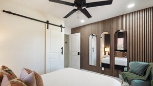 Un dormitorio con una cama, un ventilador de techo, un espejo en la pared, una mesita de noche con lámpara y una silla.