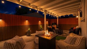 Un patio al aire libre con una fogata, zona de estar y luces colgantes.