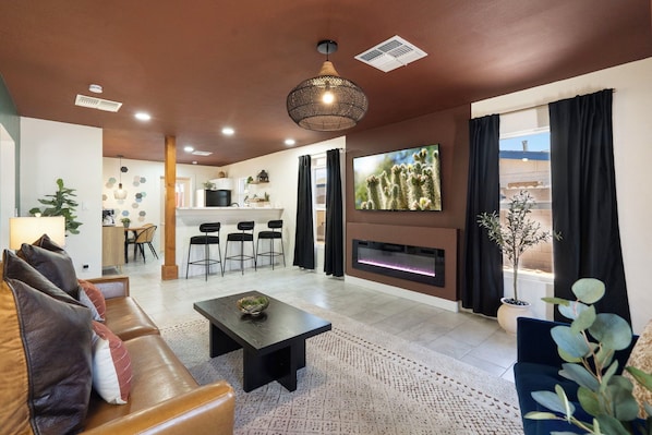 A modern living room with a sofa, coffee table, and a fireplace. There is a kitchen area in the background with bar stools.