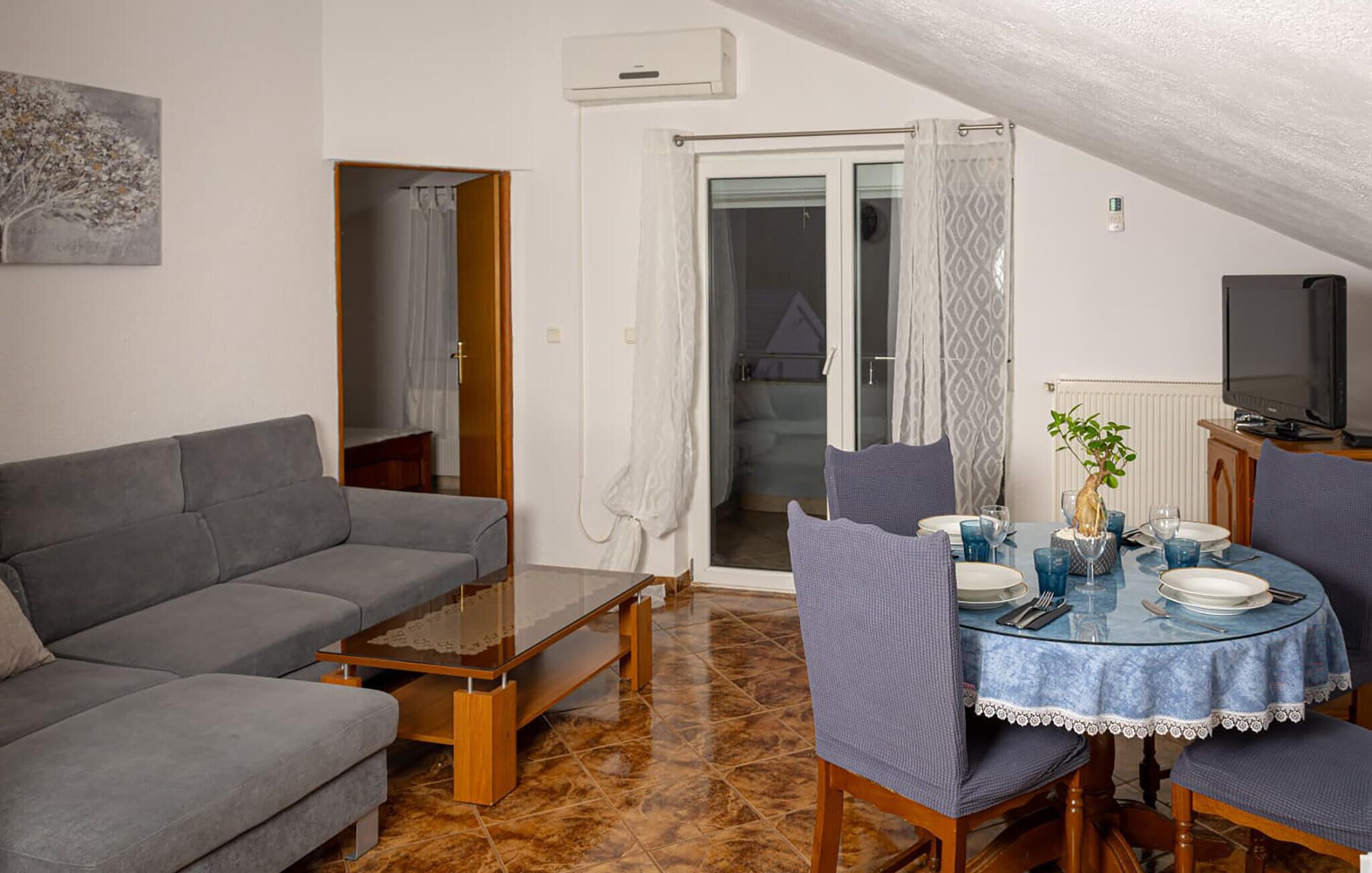 Een woonkamer met een grijze bank, een glazen salontafel en een eettafel gedekt met borden en glazen. Aan de rechterkant staat een televisie op een standaard.
