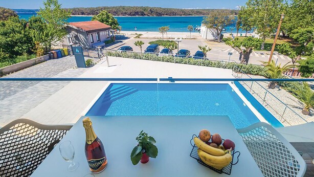 Vista a la piscina con una visión despejada del mar, una mesa con una botella, una planta y una cesta de fruta.