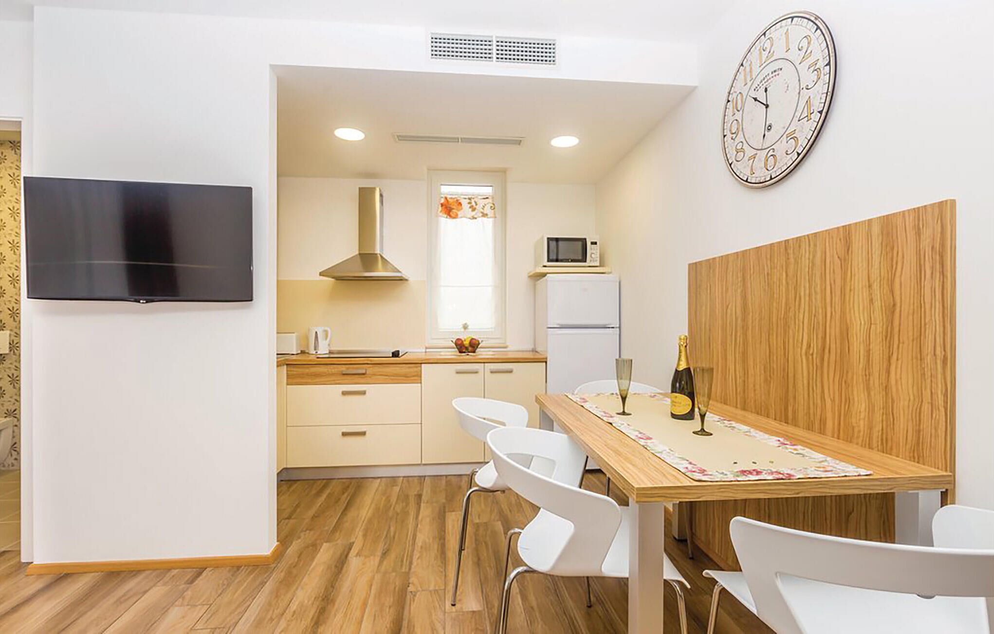 Une cuisine moderne avec des meubles blancs, une table à manger en bois avec des chaises, une horloge murale et un téléviseur à écran plat.