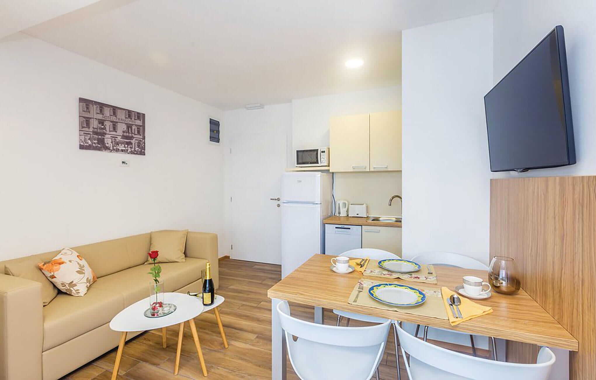 Un espacio habitable compacto con sofá, una pequeña mesa redonda con una botella y un vaso, una mesa de comedor preparada para dos personas, una zona de cocina con microondas y refrigerador, y un televisor de pantalla plana montado en la pared.