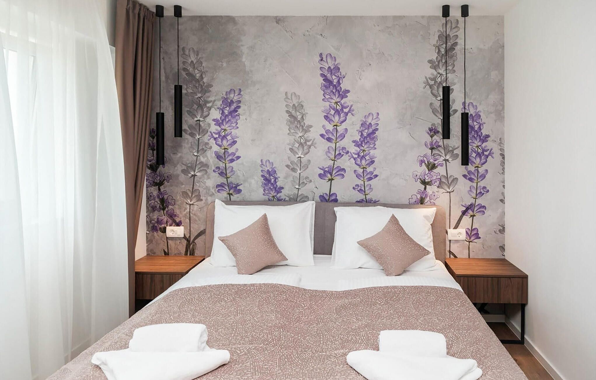 Ein modernes Schlafzimmer mit einem großen Bett, zwei Nachttischen, einer Wand mit einem floralen Wandbild und einem Fenster mit Vorhängen.
