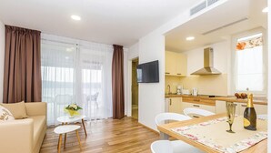 Uma sala de estar moderna com um sofá, uma mesa de jantar com cadeiras, uma pequena mesa branca com um vaso e uma área de cozinha com televisão, pia e janela com cortinas.