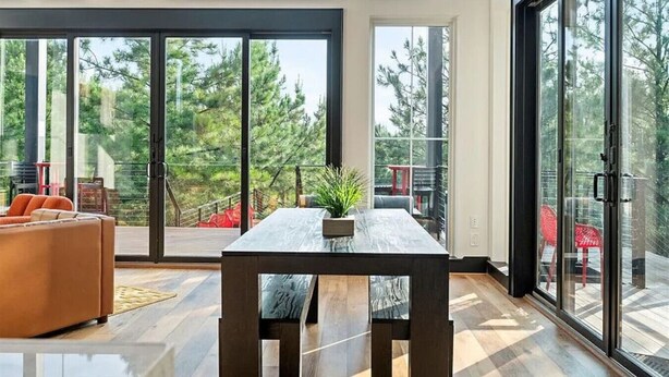 Un interior moderno con un amplio ventanal corredera que da a una terraza, una mesa de madera con una planta y un sofá.