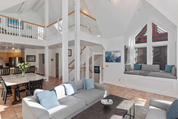 A spacious living area with a high ceiling, a dining area with a table and chairs, and a seating area with a sofa and window seat.