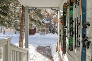 A porch with a snow-covered path leading to a house, a tree, and a wall-mounted ski rack with skis and poles.