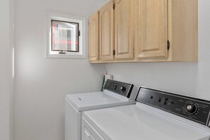 A laundry room with two washing machines, wooden cabinets above, and a window with blinds.