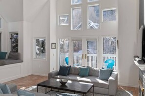 A spacious living room with a large grey sofa, a coffee table, and multiple windows offering views of a snowy outdoor area.