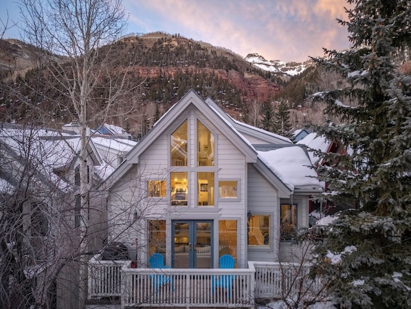 A two-story house with a gabled roof, a front porch with blue chairs, and a view of a mountainous landscape.