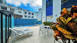 A rooftop pool area with lounge chairs and a table, surrounded by buildings with blue-tiled walls and white exteriors. - Sofistica o na Gra a Calmaria a 5 min da Barra (Salvador)
