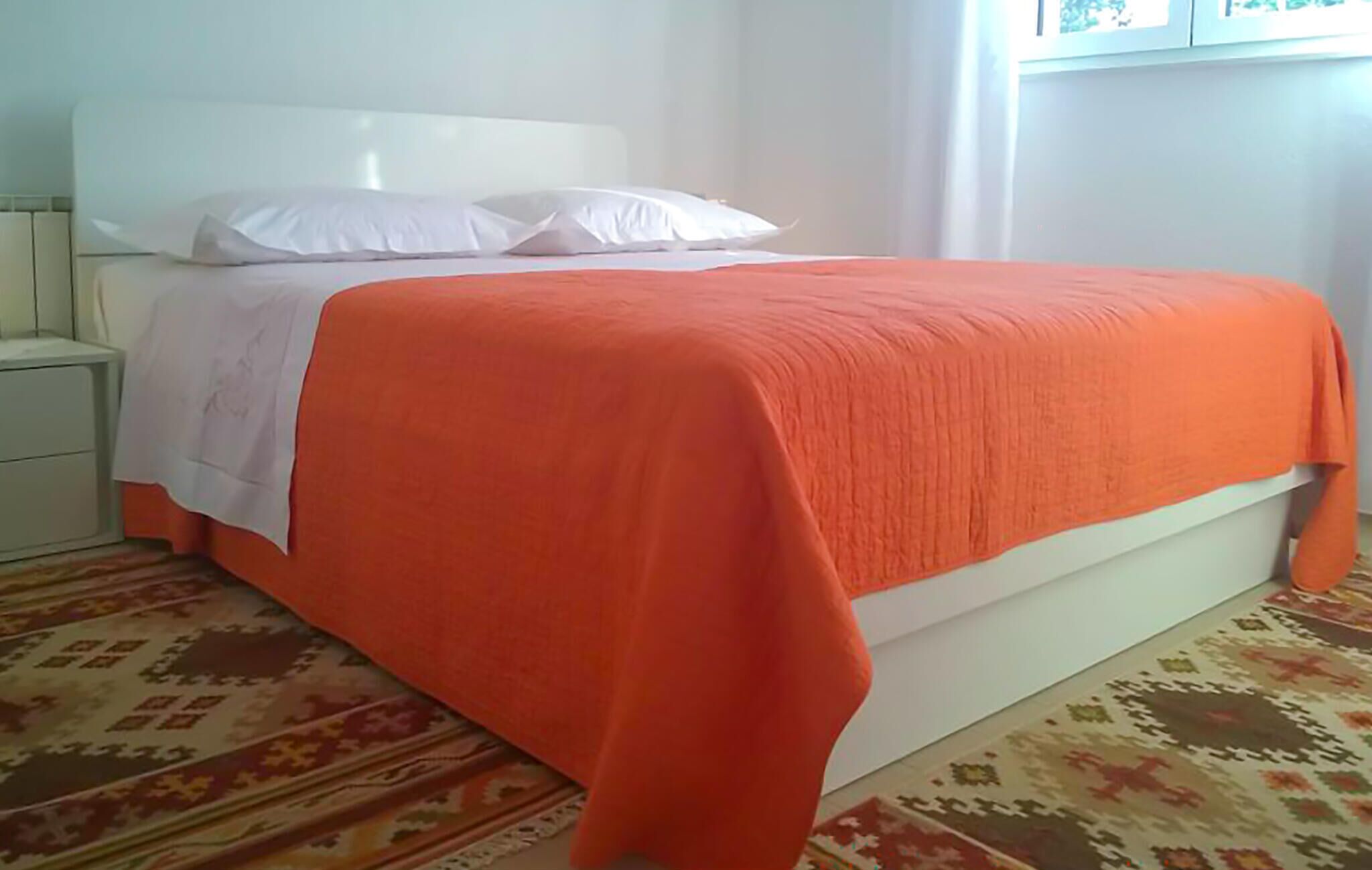 Ein Bett mit weißem Kopfteil und orangefarbener Bettdecke, ein Nachttisch und ein gemustertes Teppich.