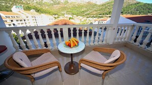 Een balkon met rieten stoelen, een klein rond tafeltje met fruit en uitzicht op een bergachtig landschap.