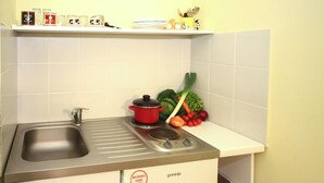 Een kleine keuken met een spoelbak, fornuis en een magnetron. Boven de spoelbak zijn planken met mokken en een koffiezetapparaat. Verse groenten, zoals wortels en tomaten, liggen op het aanrecht.