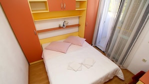 Una cama cuidadosamente tendida con almohadillas rosas y una colcha blanca, una lámpara de noche y suelo de madera.
