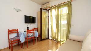 Una habitación pequeña y bien iluminada, con piso de madera, un sofá, una mesa de comedor con dos sillas, un televisor montado en la pared y una puerta corrediza de vidrio con cortinas.